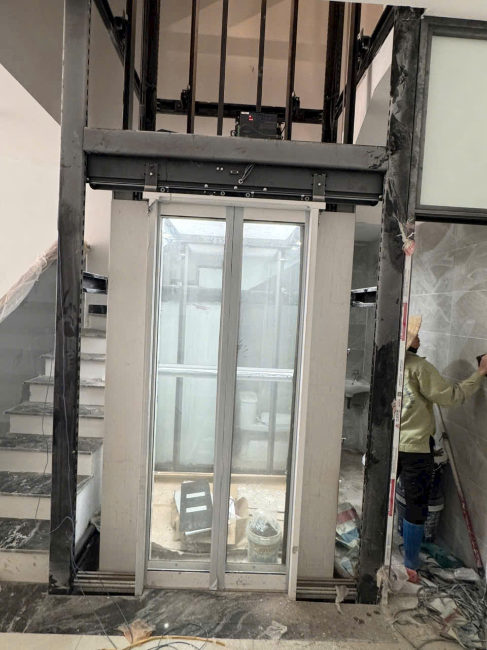 Thang máy gia đình cửa kính đang lắp đặt giữa hai nhánh cầu thang trong nhà xây dựng